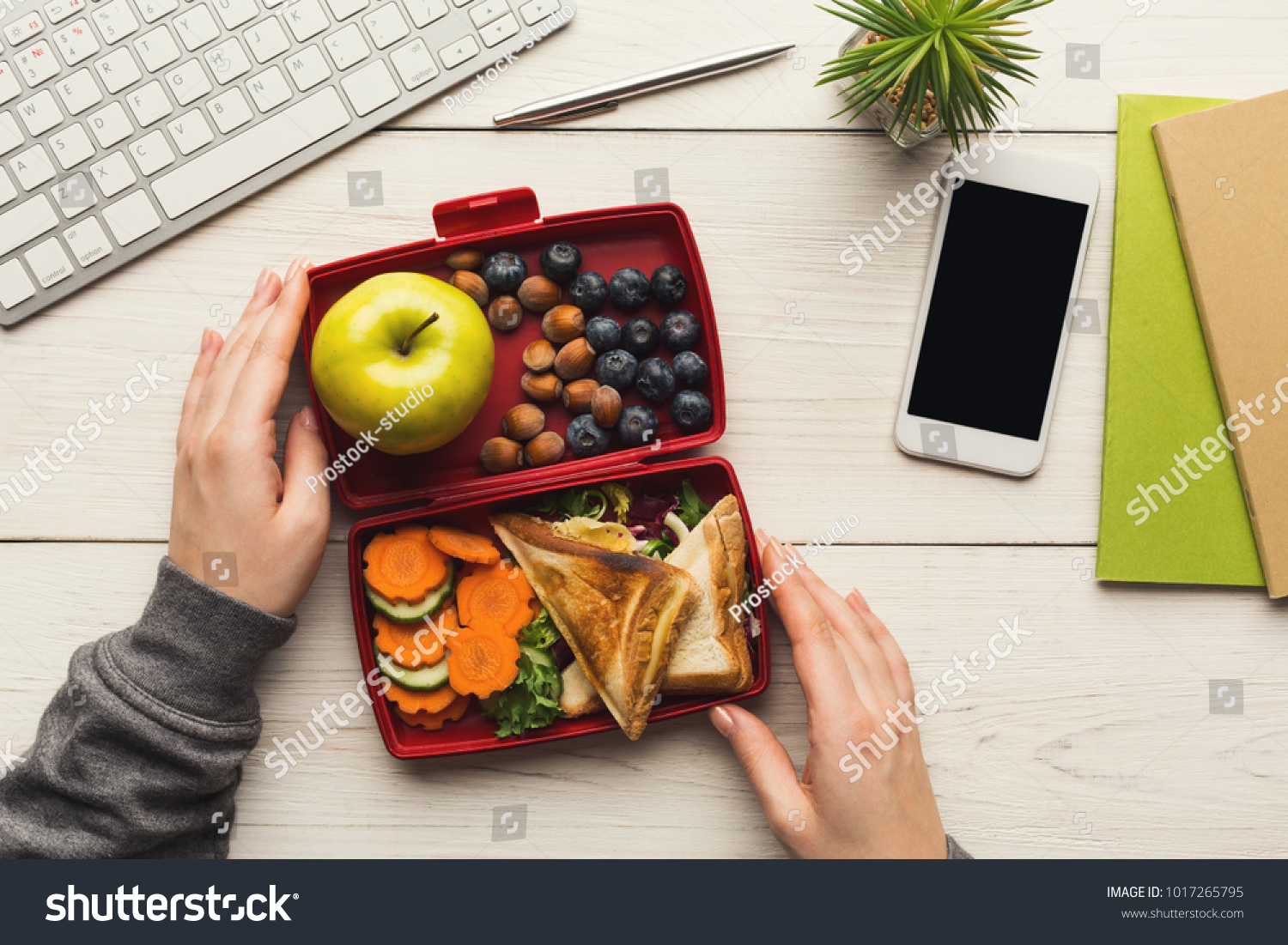 Healthy snack at office workplace. Businesswoman eating organic vegan meals from take away lunch box at wooden working table with computer keyboard and smartphone with empty screen for copy space #1017265795