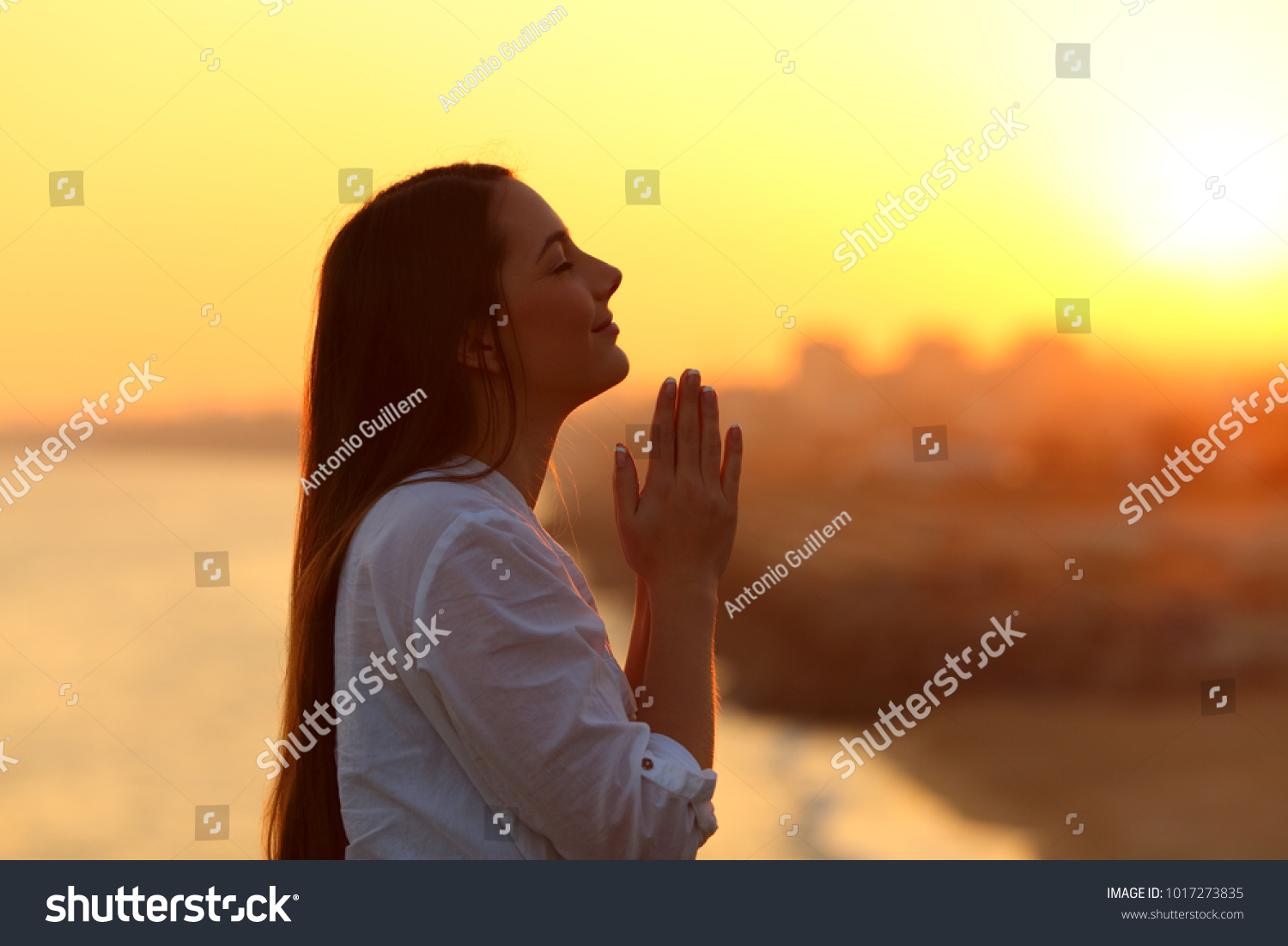 Side view backlight portrait of a woman praying and looking above at sunset #1017273835