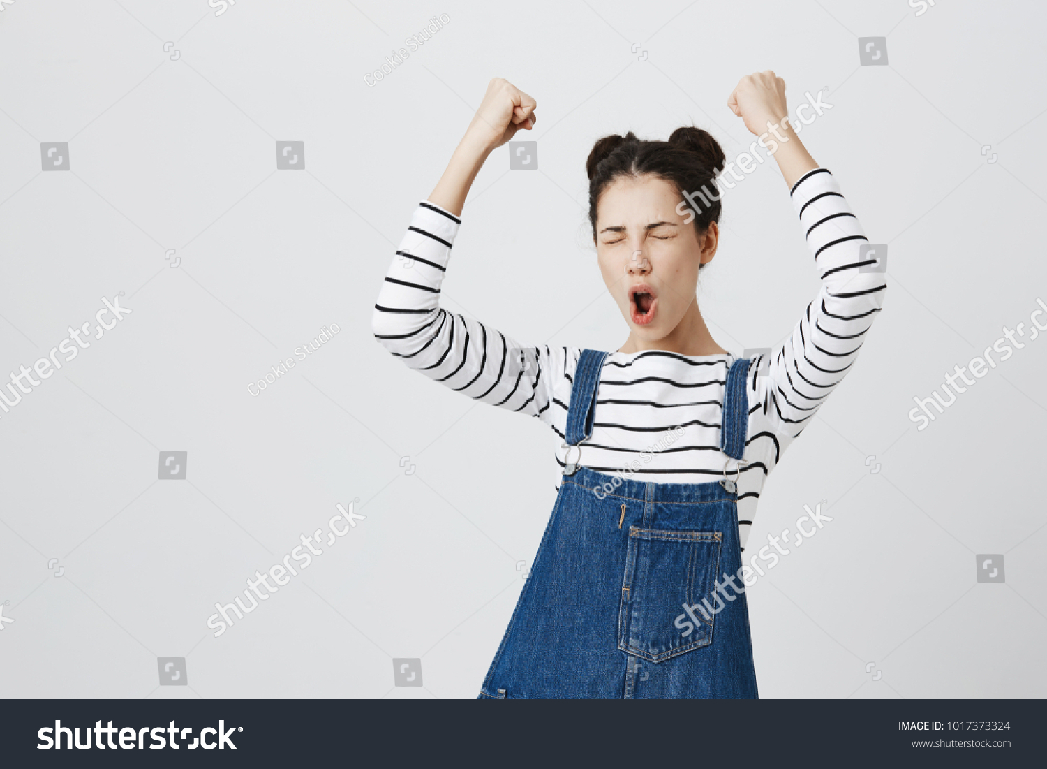 Brunette girl with hairbuns in striped top excited and glad to achieve victory, clenches fists, screams in excitement with closed eyes, happy to pass all exams at university, successful person. #1017373324