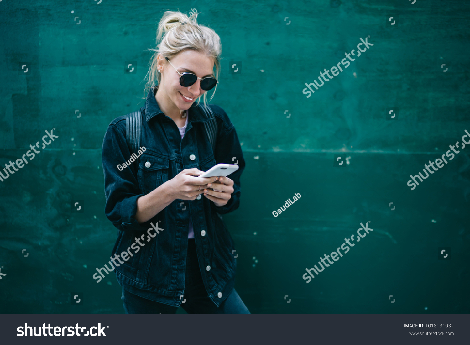 Smiling caucasian young woman sending message on smartphone standing near promotional copy space background, positive trendy hipster girl blogging in social network using mobile phone with internet #1018031032