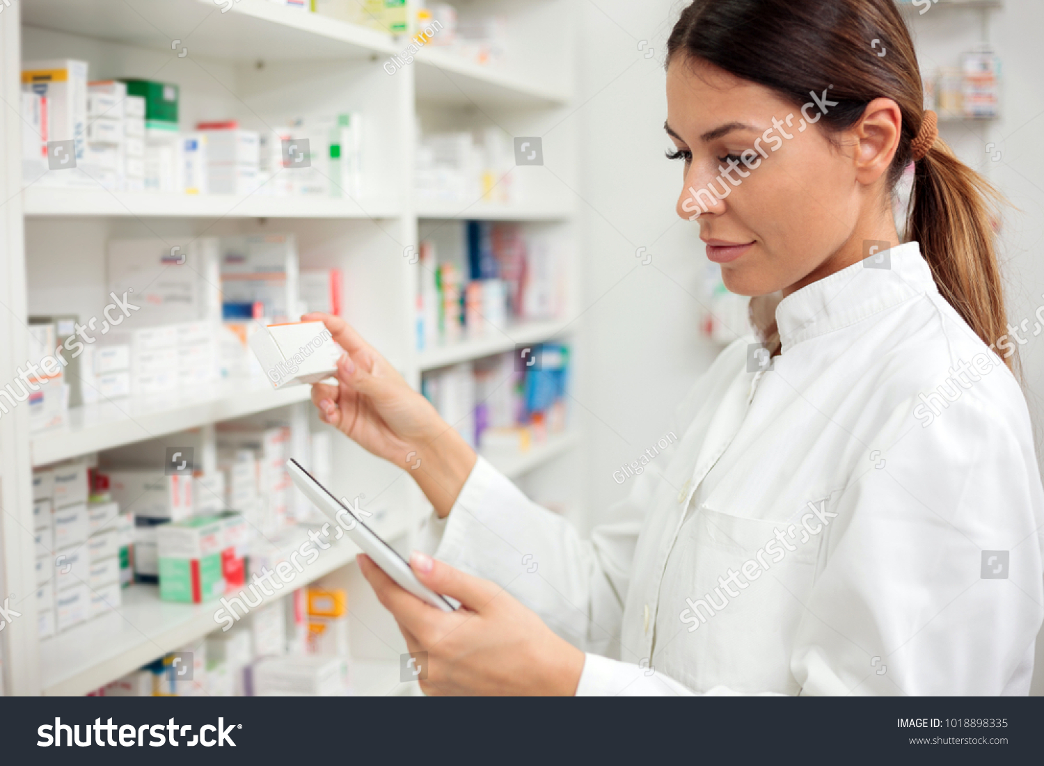 Medicine, pharmaceutics, health care and people concept - Serious young female pharmacist taking medications from the shelf. #1018898335