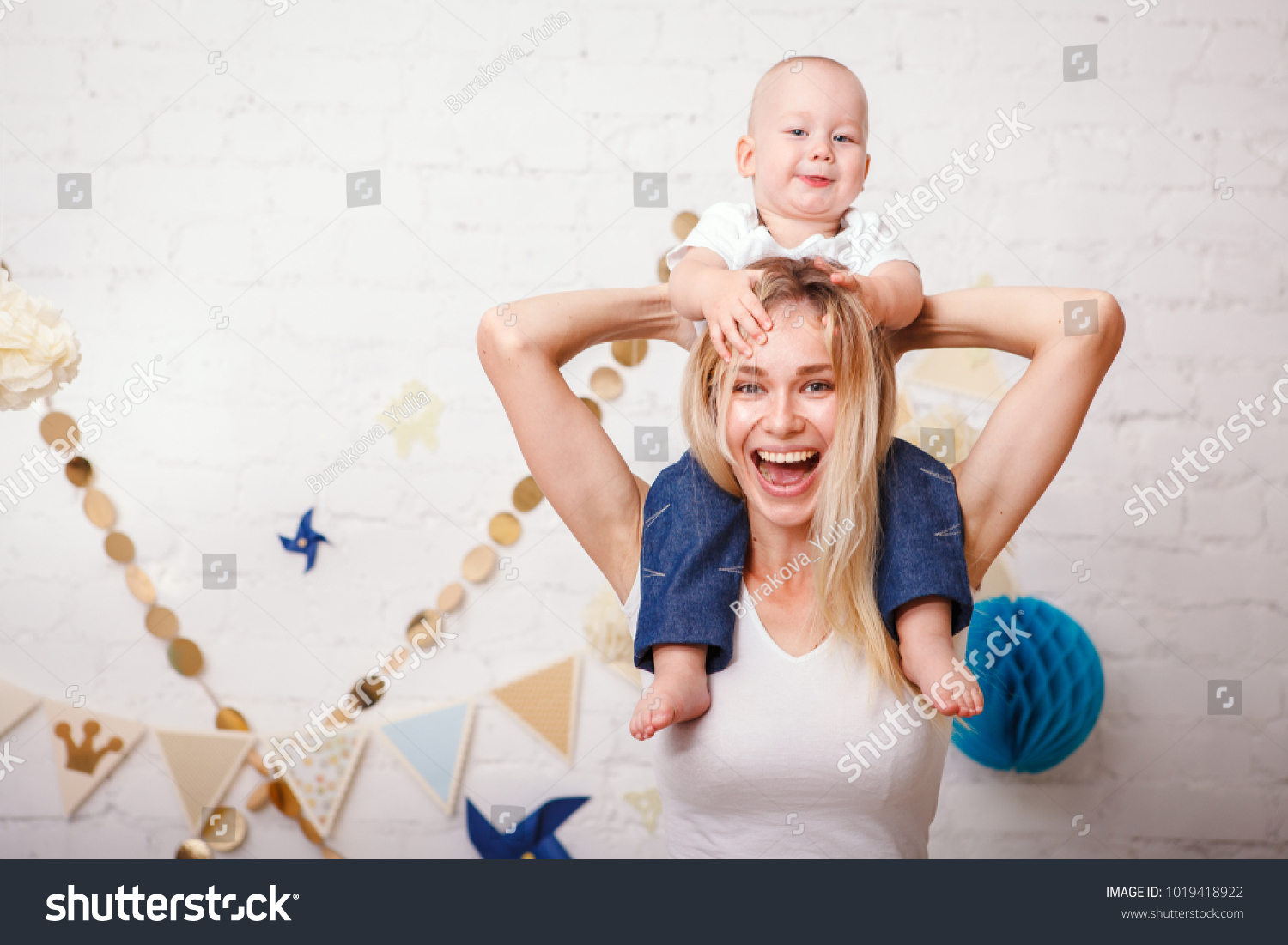 A cute handsome one-year-old baby sitting by her mother's neck grabs her by the hair and laughs. A beautiful young mother with her son play laughing and look at the camera. Mothers Day. #1019418922
