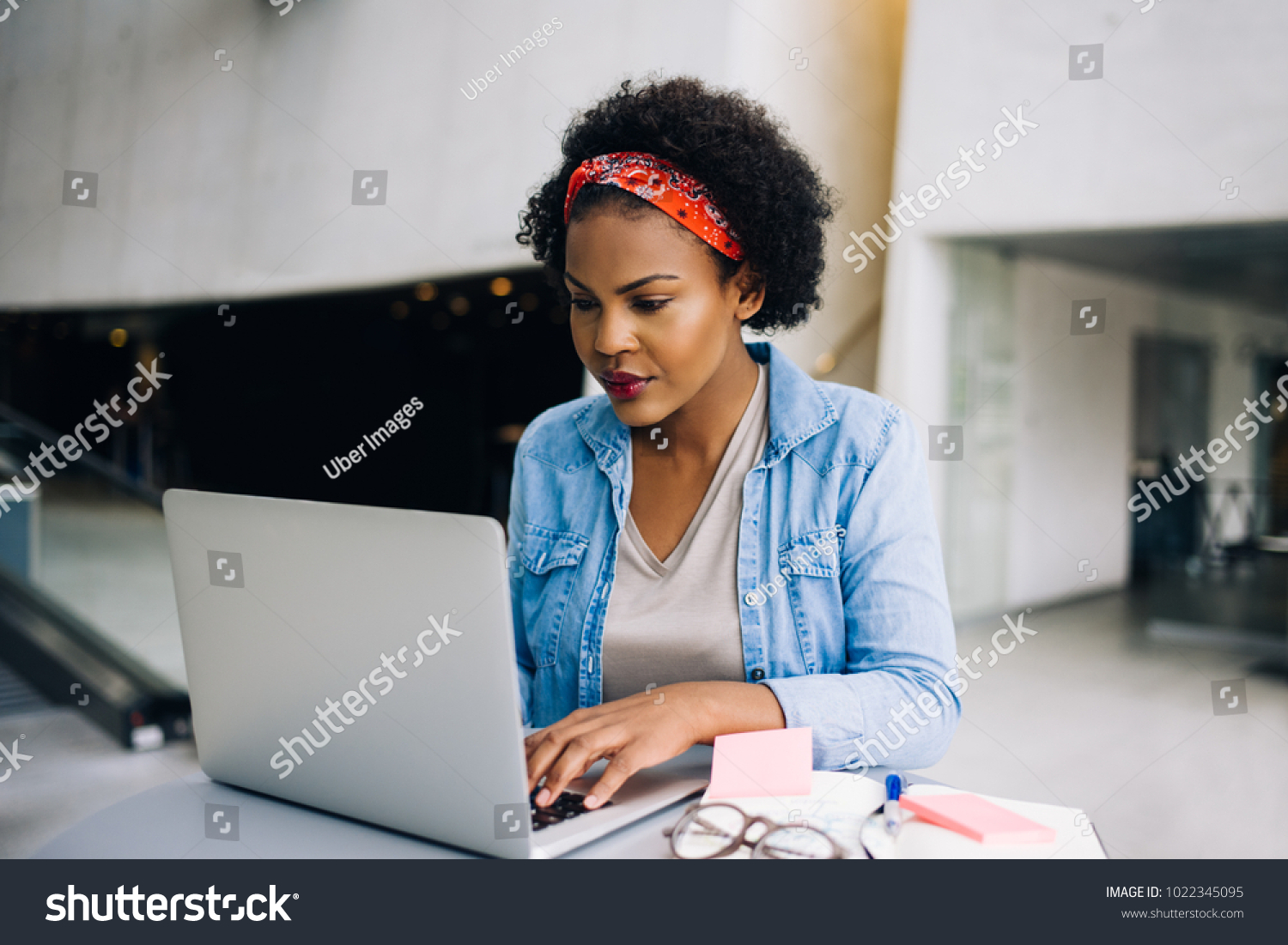 Focused young African female entrepreneur hard at work on a laptop while sitting at a table in the lobby of a modern office building #1022345095