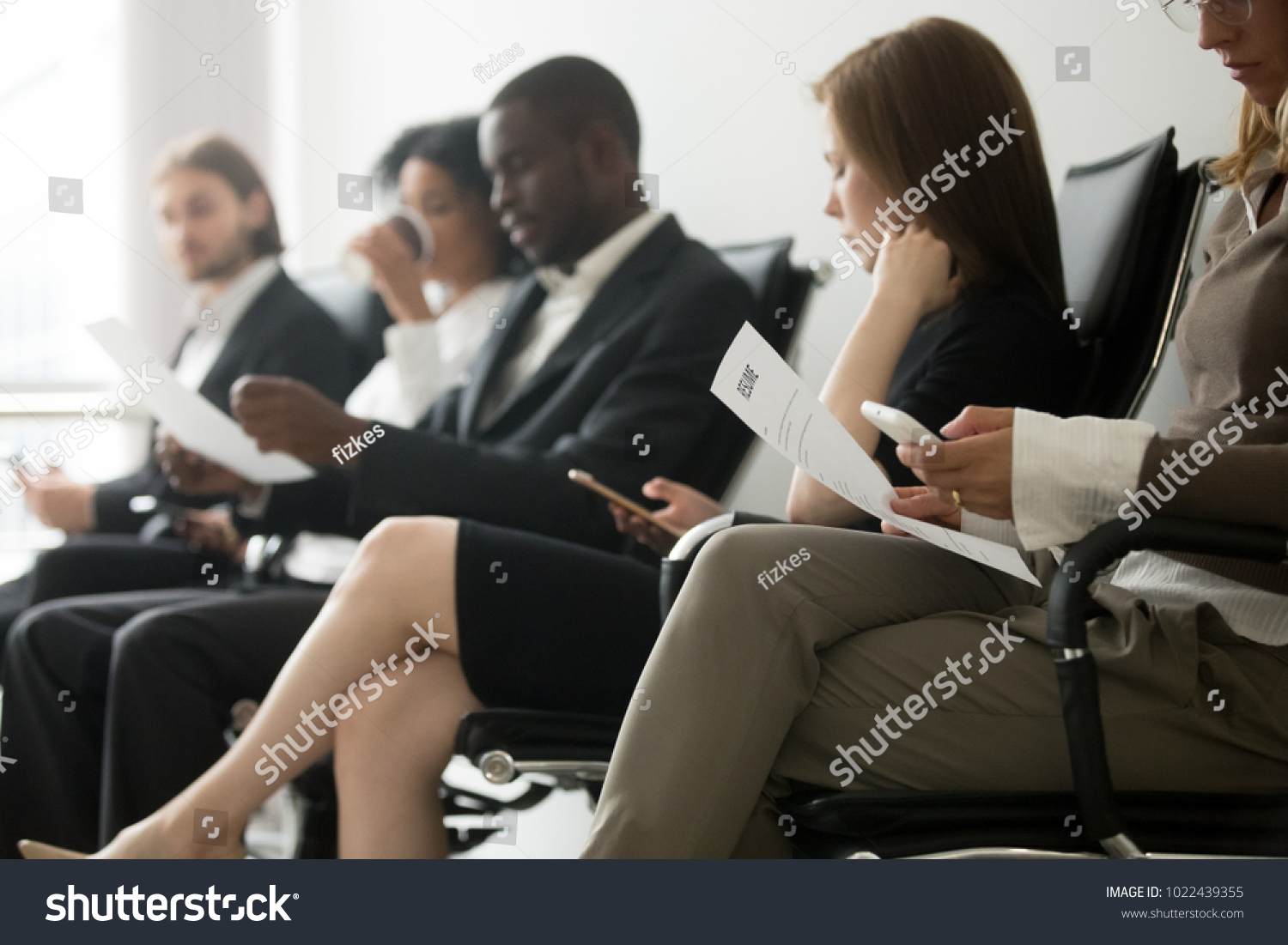Multi-ethnic applicants sitting in queue preparing for interview, black and white vacancy candidates waiting on chairs holding resume using smartphones, human resources, hiring and job search concept #1022439355
