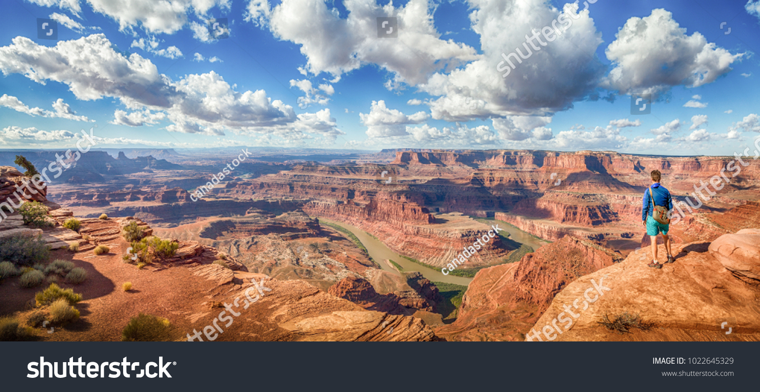 Panoramic view of young hiker standing on a cliff in scenic Dead Horse Point State Park enjoying the view on a beautiful sunny day with blue sky and dramatic clouds in summer, Utah, USA #1022645329
