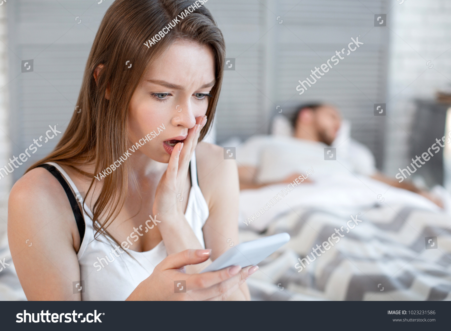 Oh my god. Pretty startled heart-broken woman reading her mans messages while he sleeping and she sitting on the bed #1023231586