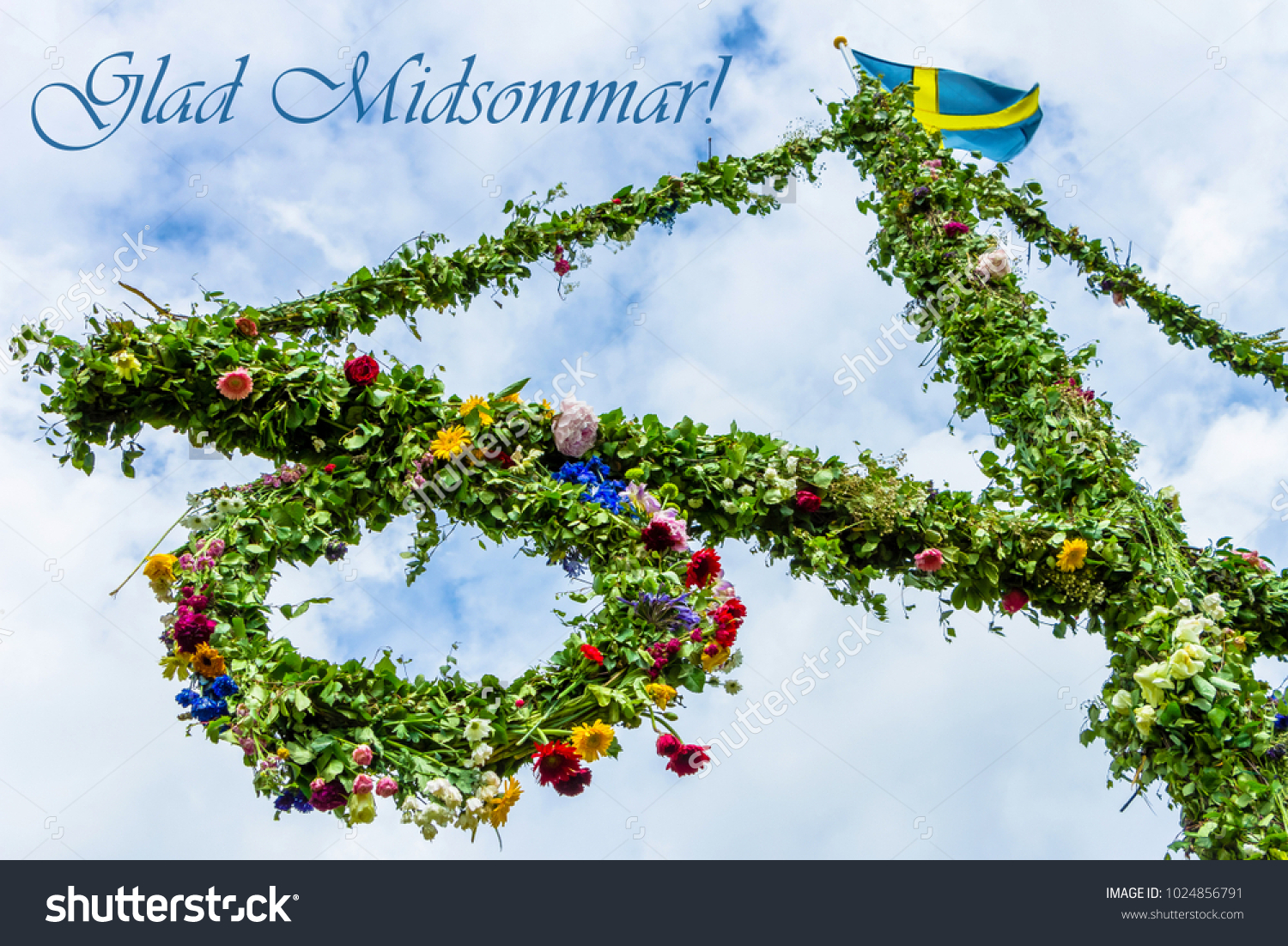 A pole and flag against blue sky and white clouds. A maypole decorated, covered in flowers and leaves. Pole after celebrating midsummer. Midsummer traditional Swedish symbol.  Kort Glad Midsommar. #1024856791
