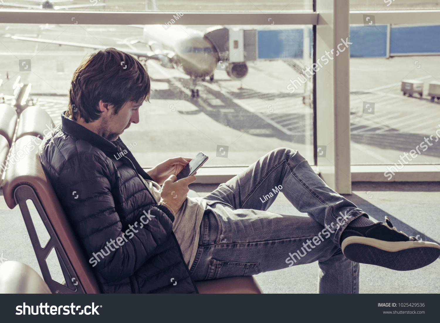 Young caucasian man in jeans and outerwear sitting in chair airport waiting hall and using smartphone. Online remote banking, hotel booking, mobility, reading news, use of 3 G and 4 G network. #1025429536