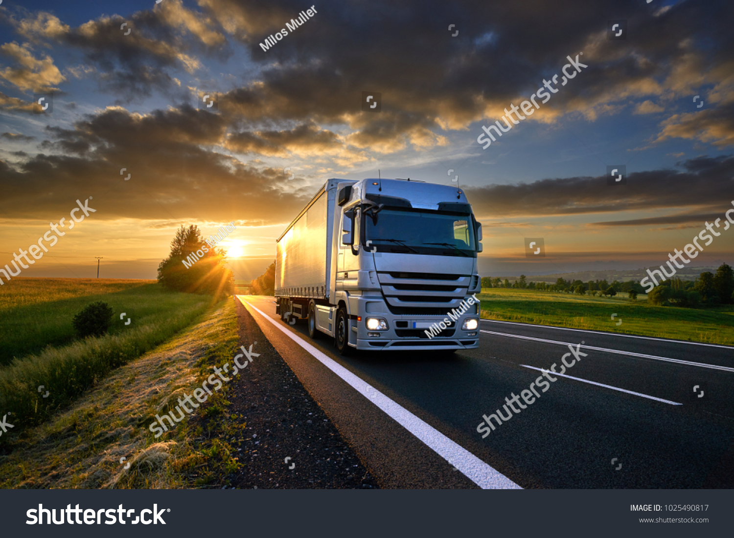 Truck driving on the asphalt road in rural landscape at sunset with dark clouds #1025490817