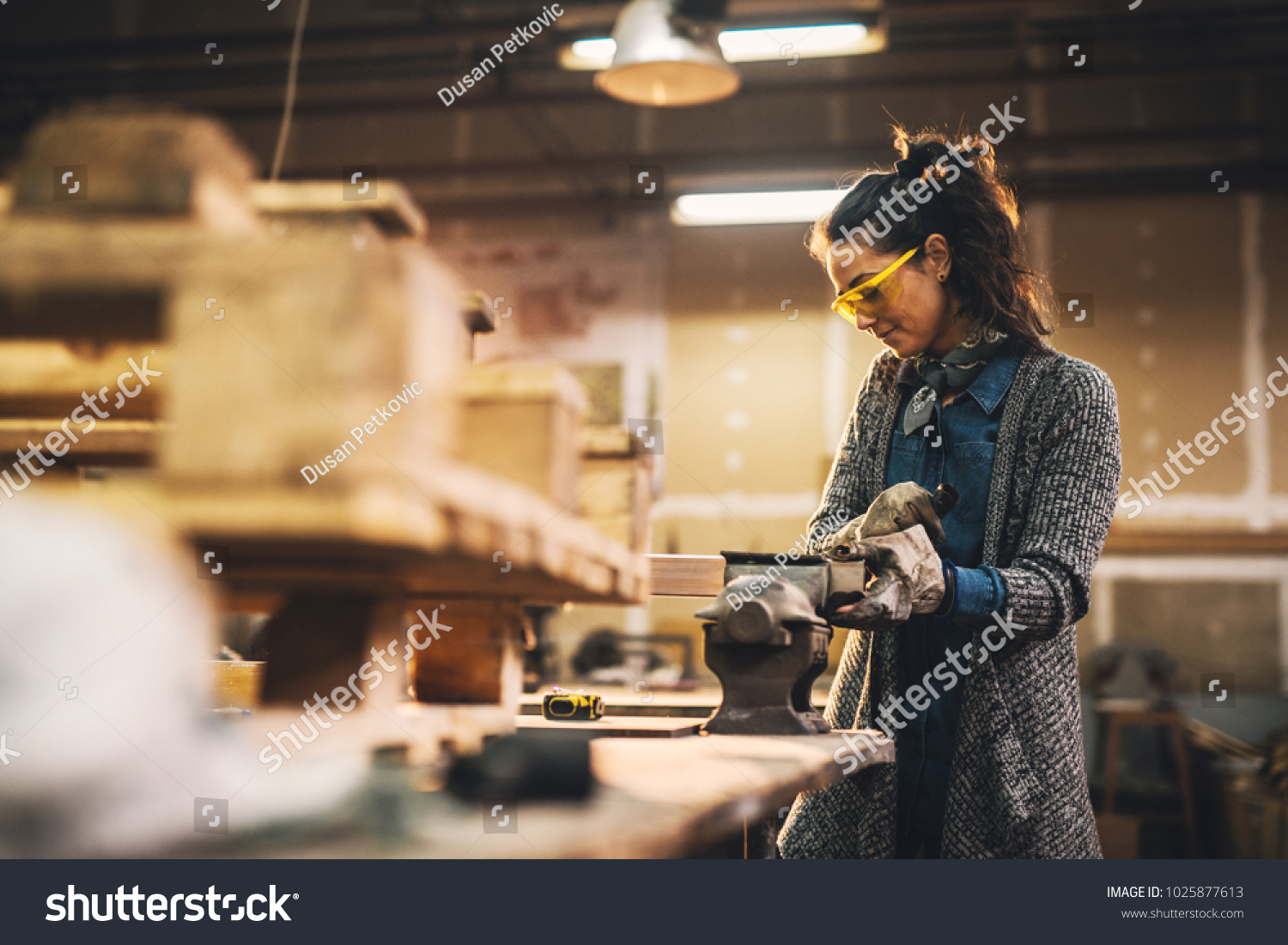 Portrait view of satisfied smiling middle aged professional female carpentry worker with steel vise on the table in the workshop. #1025877613