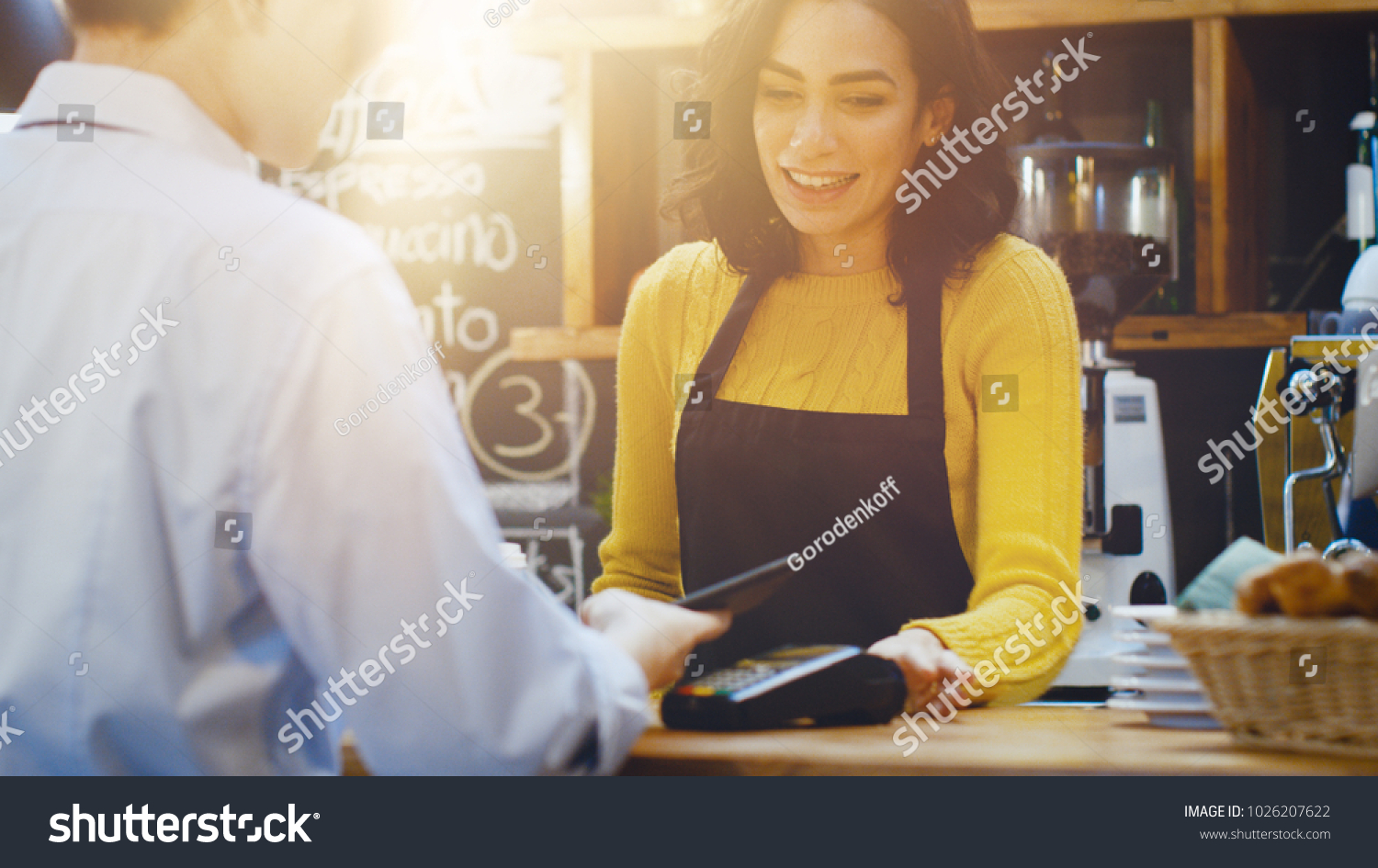 In the Cafe Beautiful Hispanic Woman Makes Takeaway Coffee For a Customer Who Pays by Contactless Mobile Phone to Credit Card System. #1026207622