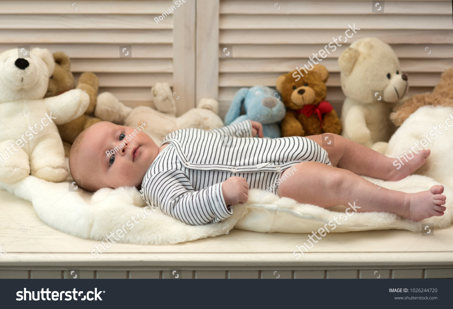 Baby boy in striped bodysuit. Baby lying on soft white duvet. Childhood and innocence concept. Infant with blue eyes and peaceful smile among teddy bears on wooden background, defocused #1026244720