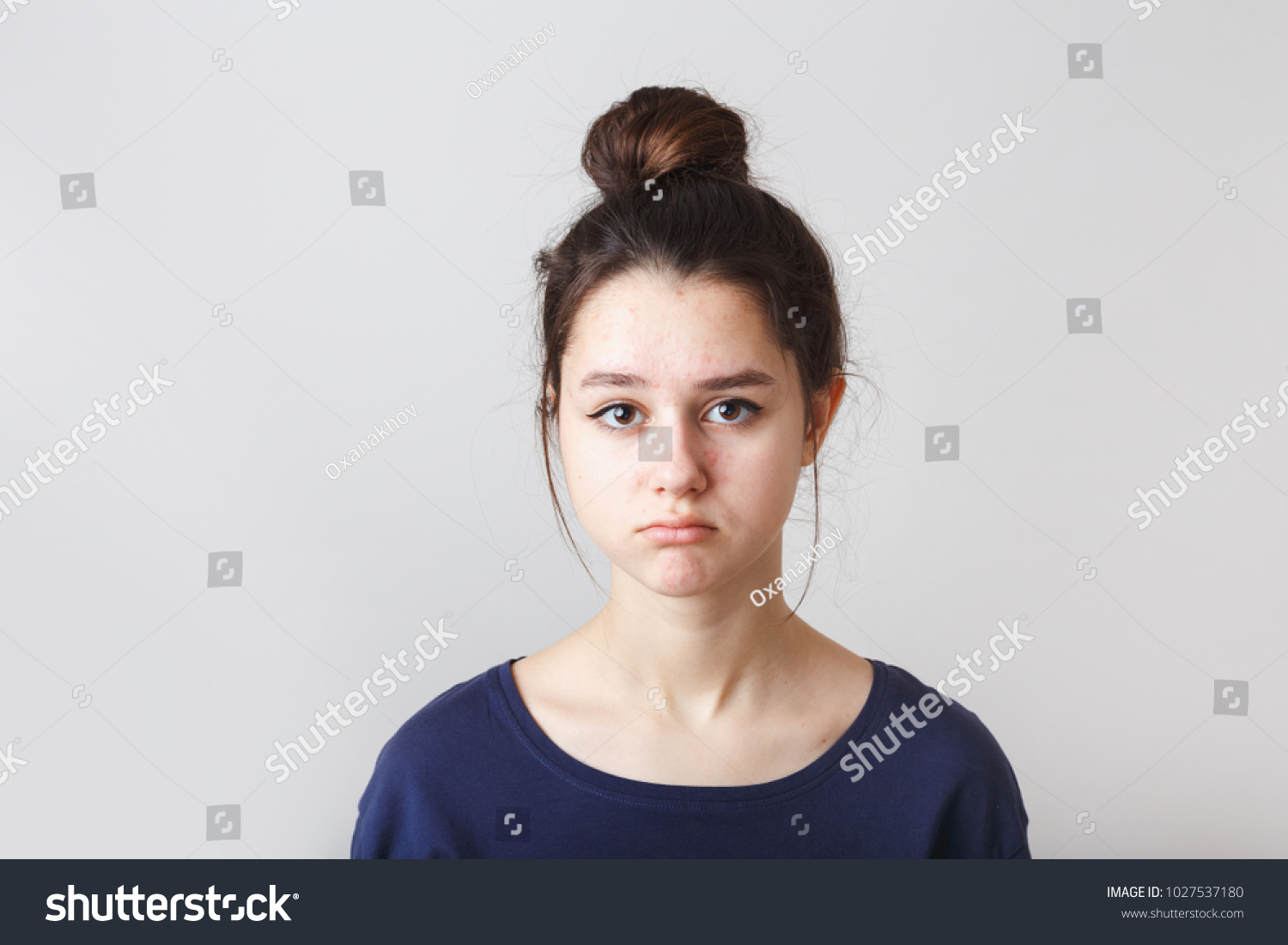 portrait of a pimply teenage girl in a blue T-shirt on a gray background, sad face #1027537180