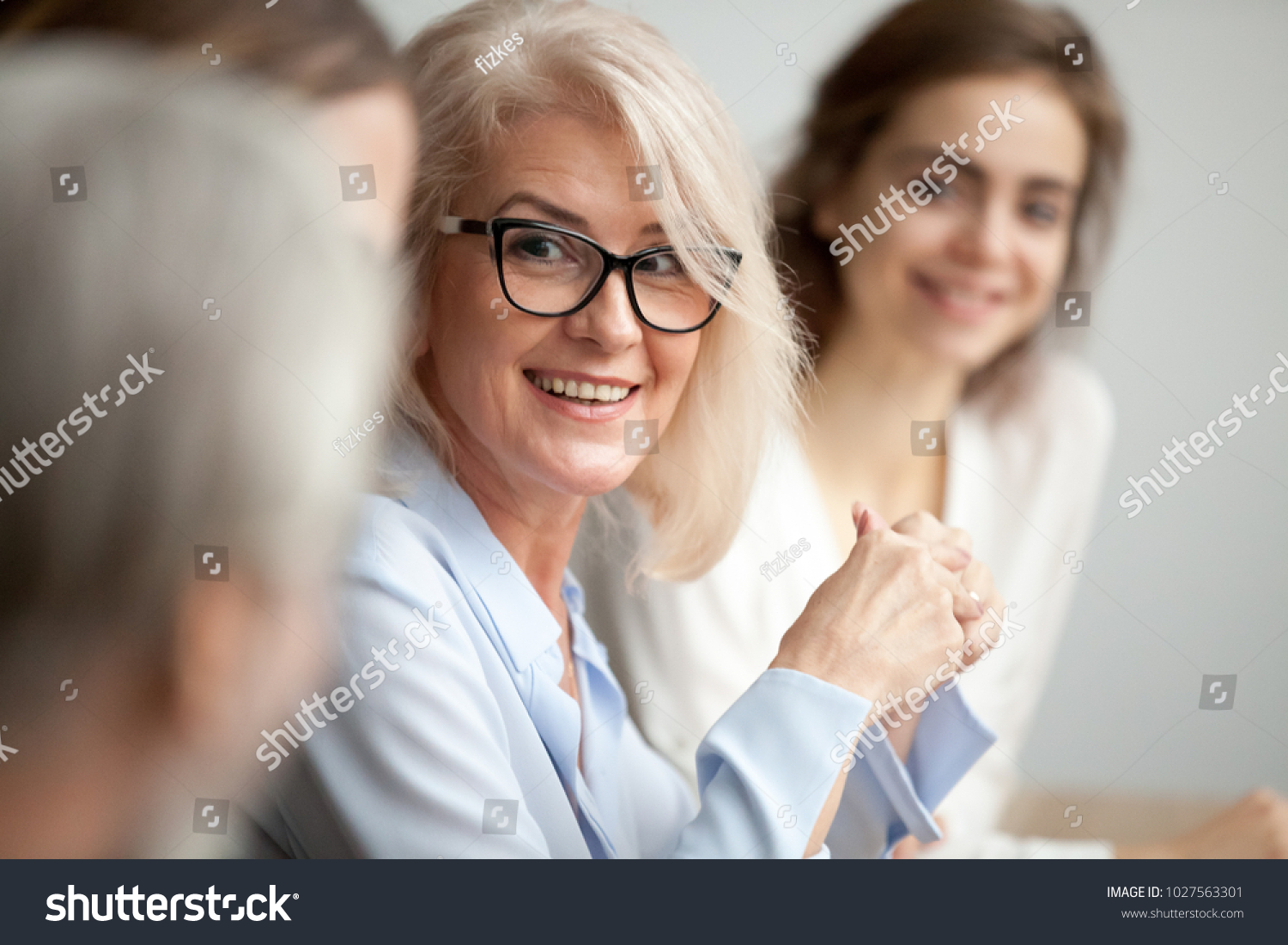 Smiling aged businesswoman in glasses looking at colleague at team meeting, happy attentive female team leader listening to new project idea, coach mentor teacher excited by interesting discussion #1027563301