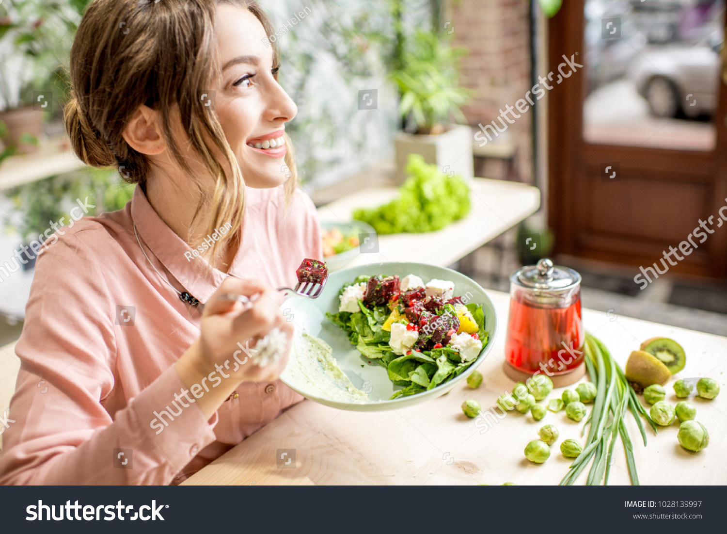 Young woman eating healthy food sitting in the beautiful interior with green flowers on the background #1028139997
