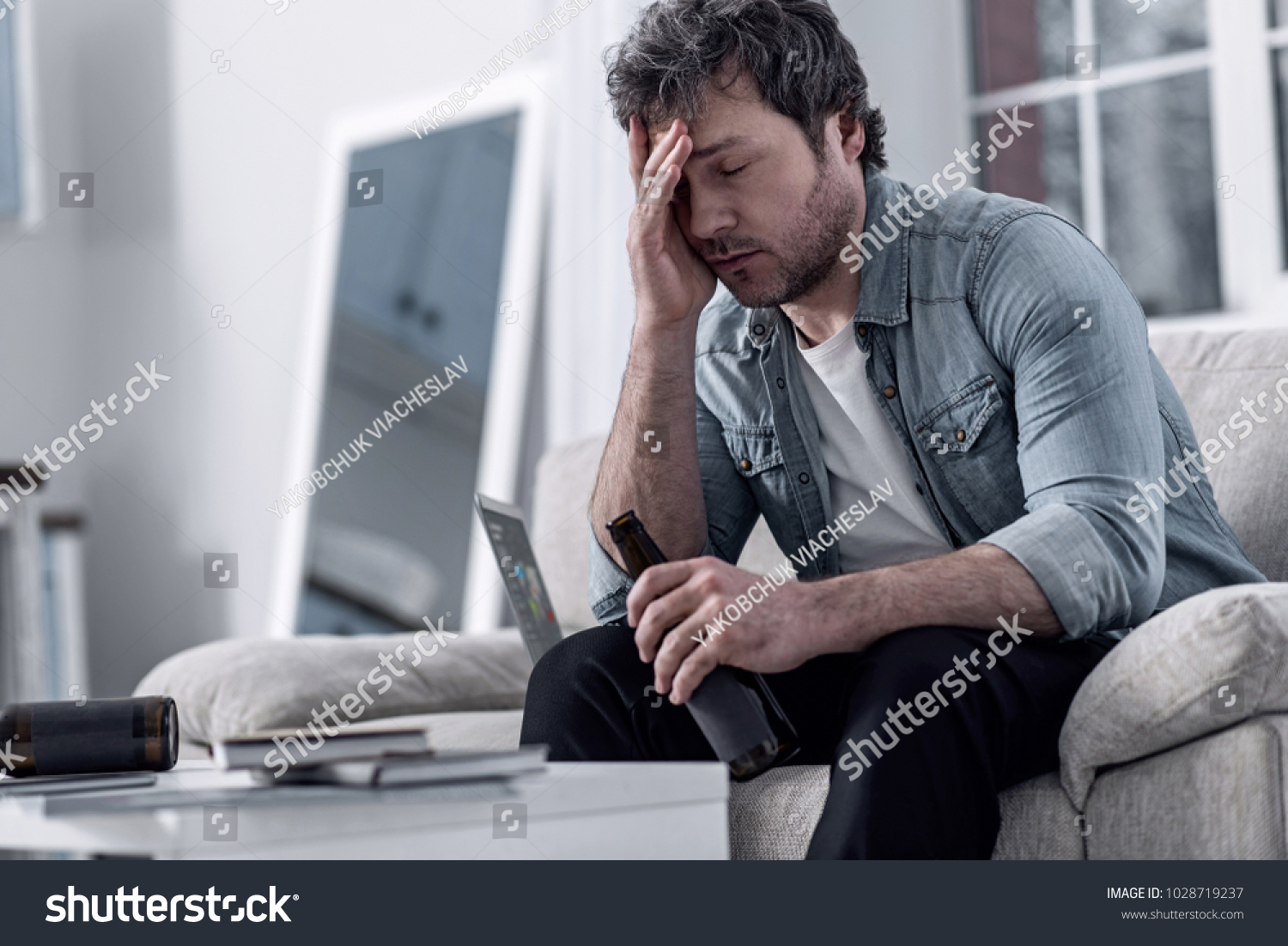Awful feeling. Unhappy depressed man sitting with his eyes closed and touching his forehead while sitting with a bottle in his hand and feeling bad after drinking too much alcohol #1028719237