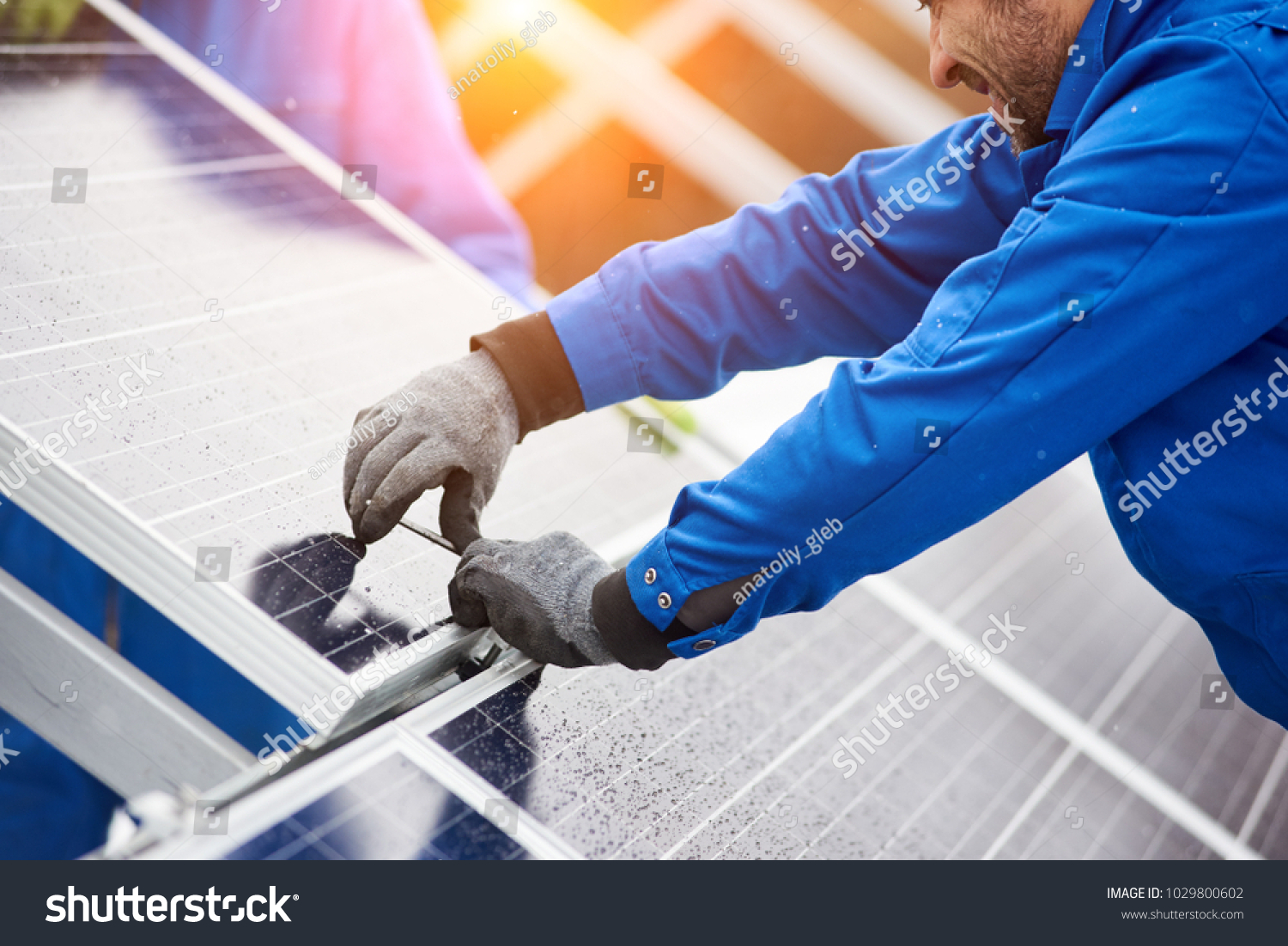 Smiling male technician in blue suit installing photovoltaic blue solar modules with screw. Man electrician panel sun sustainable resources renewable energy source alternative innovation #1029800602