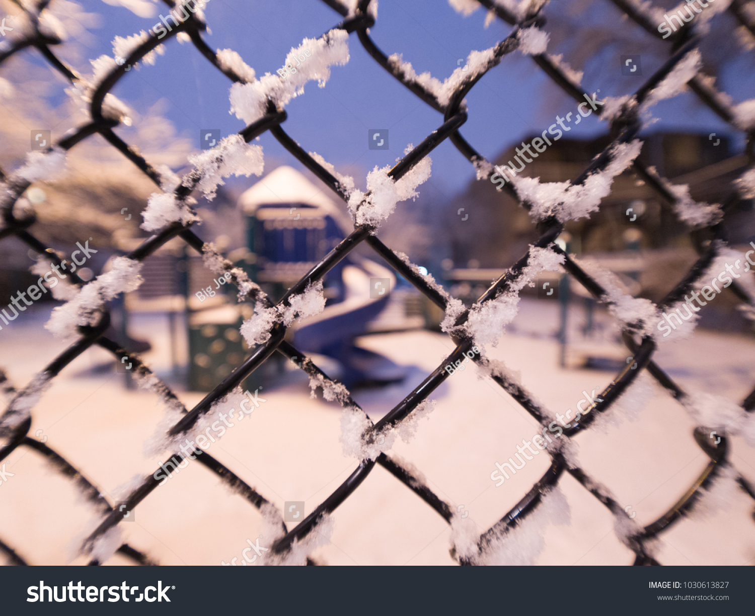 View looking through a snow covered chain link fence to a playground slide on a cold quiet evening in winter in Chicago with blue sky beyond. #1030613827