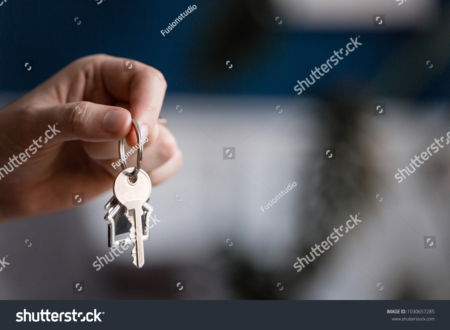 Mortgage concept. Men hand holding key with house shaped keychain. Modern light lobby interior. Real estate, moving home or renting property. #1030657285