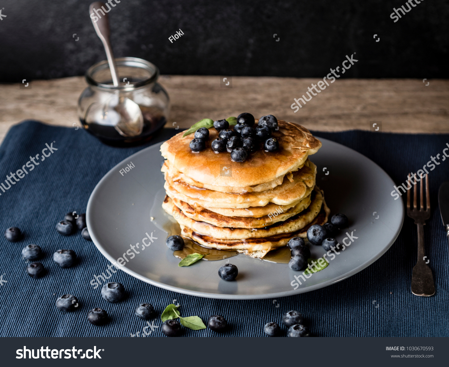 image shows a homemade fluffy pancake with blueberry on the top; situation is decorated with rustiv wooden table, placemat, silver cutlery, honey glass #1030670593