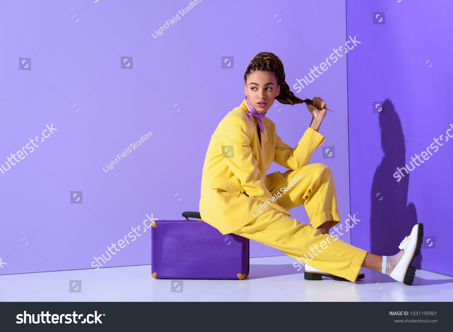 african american girl posing in yellow suit sitting on purple suitcase, on trendy ultra violet background #1031199901