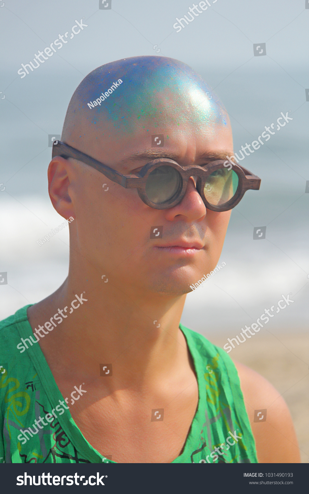 A bald and unusual young man, a freak, with a shiny bald head and round wooden glasses on the background of the beach and the sea. Humor and eccentricity. Unusual appearance. Humorist. #1031490193
