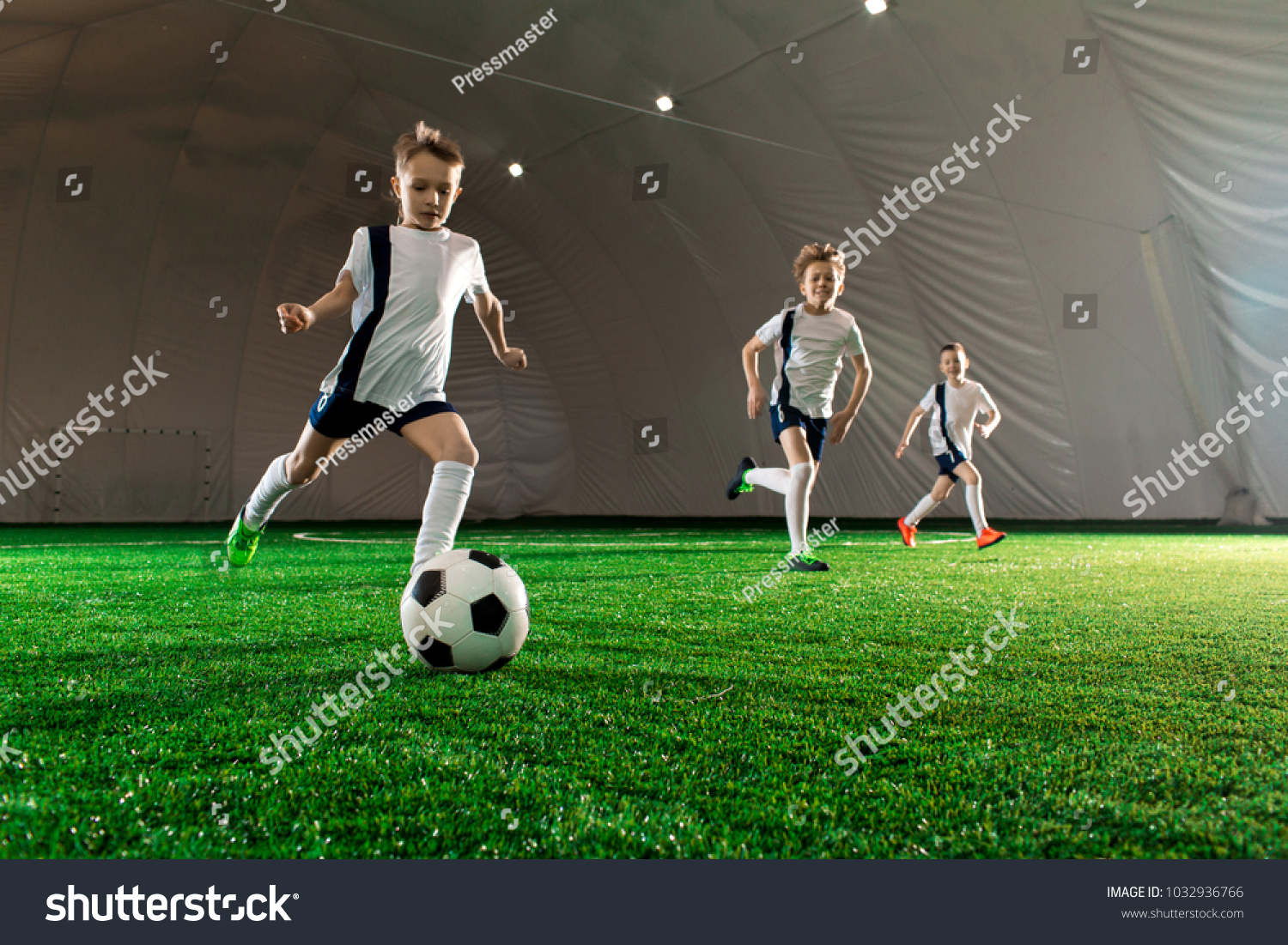 Three boys in uniform running after ball down green field while training indoors #1032936766