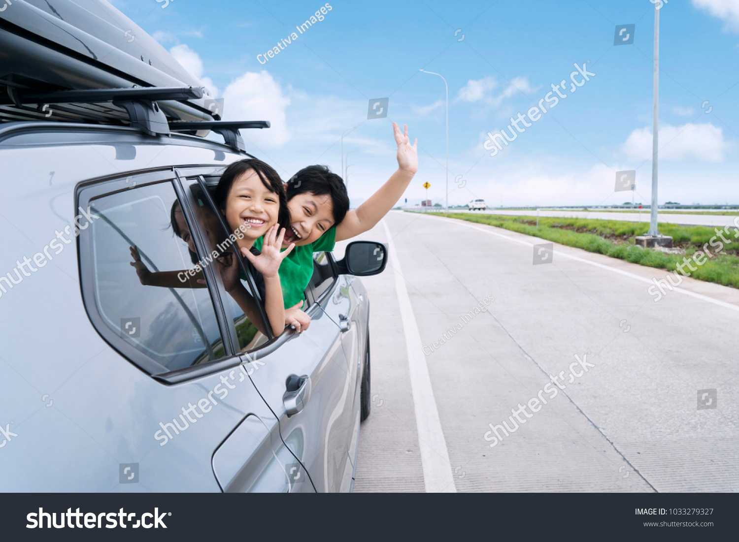 Happy siblings waving hands travel by car against blue sky. Summer road trip concept #1033279327