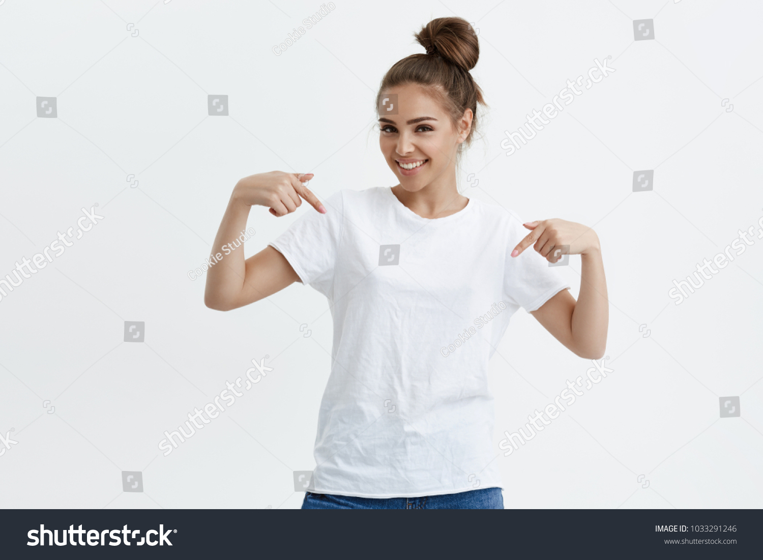 Charming emotive caucasian woman pointing down or at her t-shirt while smiling joyfully and expressing positive emotions over white background. Girl trained a lot, she wants to show her muscles #1033291246