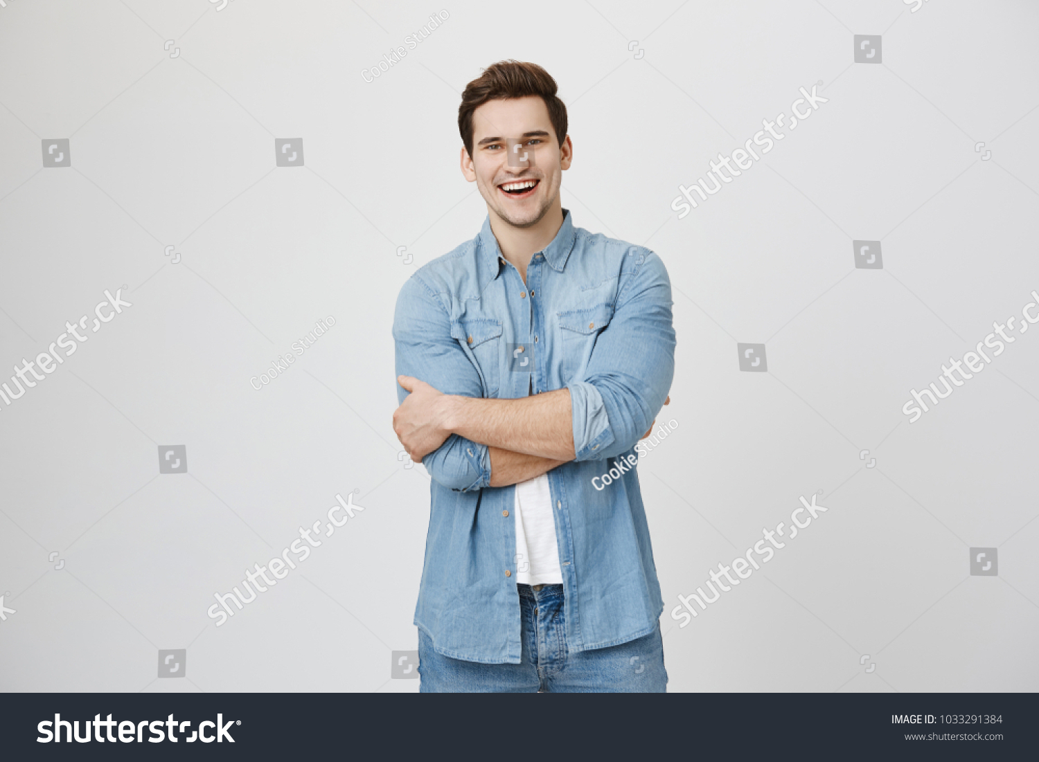 Portrait of guy wearing denim shirt, standing with crossed hands while laughing, isolated over white background. Attractive hairdresser greets new visitor offering him cup of coffee #1033291384