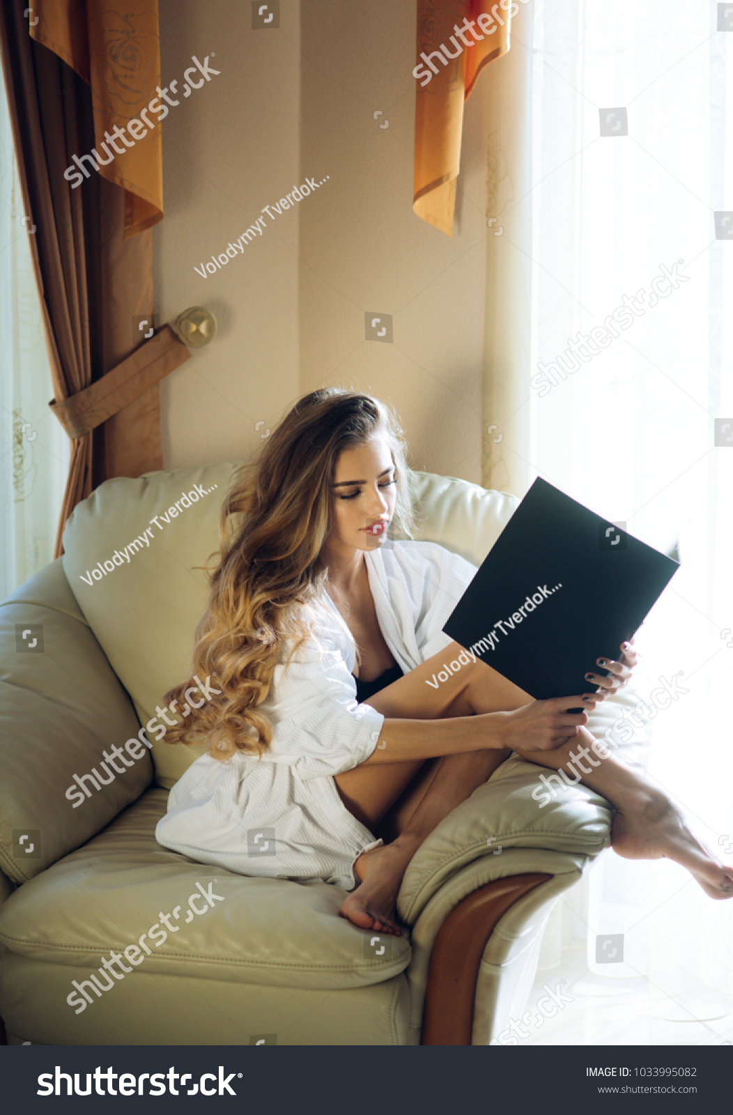 Lady with busy face in bathrobe reads book near window. Girl enjoys morning reading siting on armchair. Sexy nude woman with long hair looks hot and desire. Attraction and desire concept. #1033995082