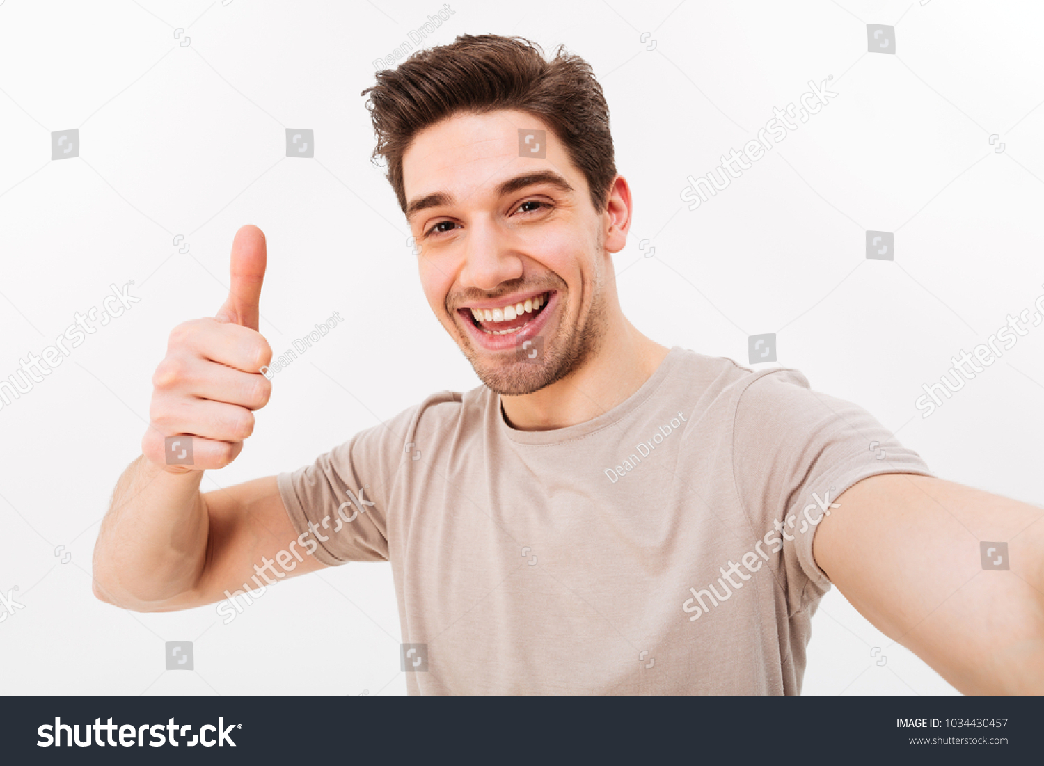 Photo of handsome man in casual t-shirt and bristle on face smiling on camera with thumb up while taking selfie isolated over white background #1034430457