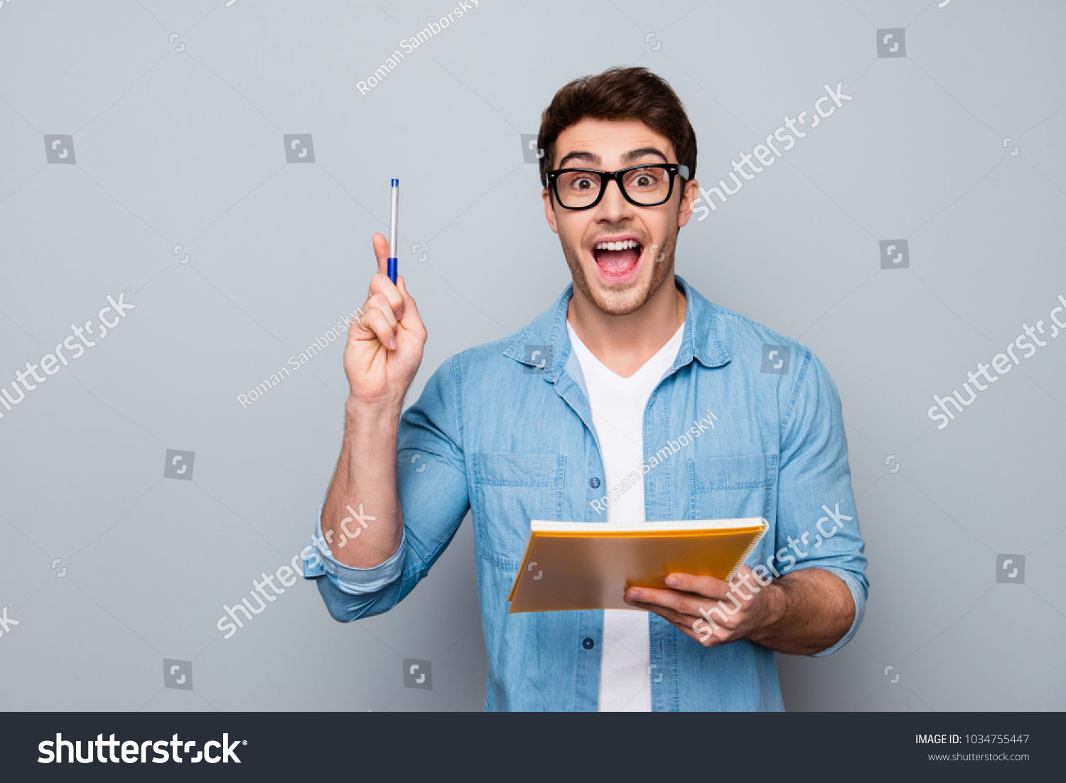 Handsome, attractive, glad, positive, funny guy in glasses with wide open mouth finally found a solution how to make exercise, having raised pen and notebook in hands, isolated on grey background #1034755447
