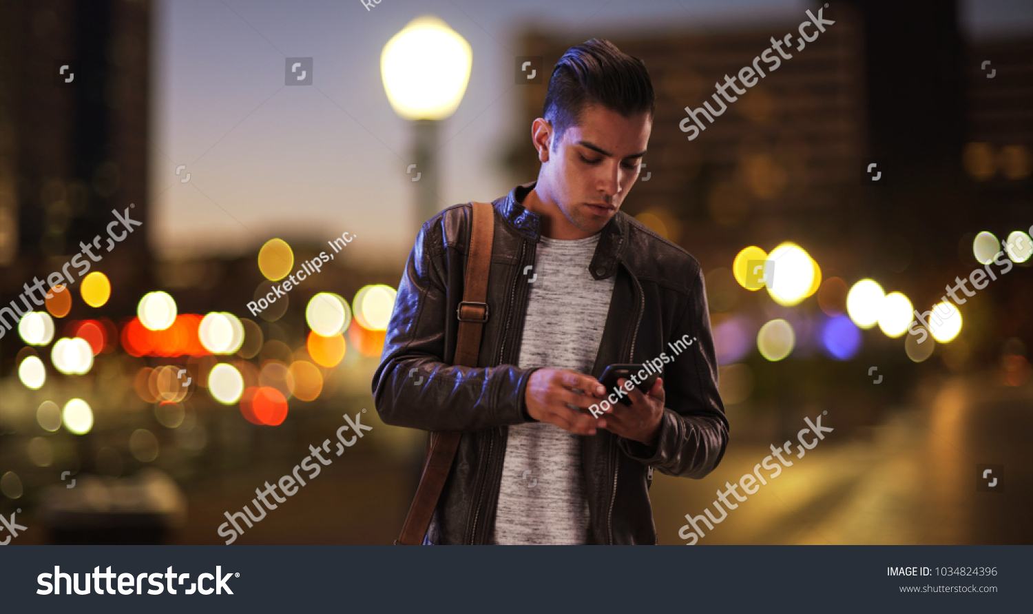 Millennial Hispanic man standing on city bridge at night looking at smartphone #1034824396