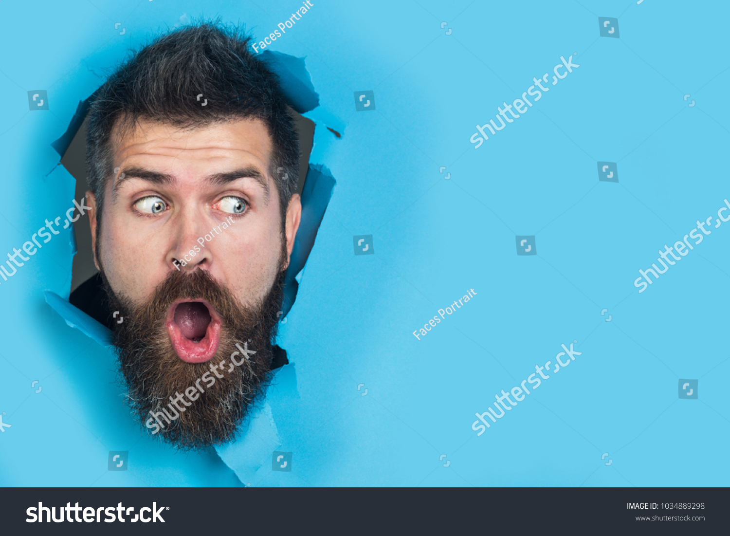 View of male face through hole in blue paper. Surprised bearded man making hole in paper. Copy space for advertising, to insert text or slogan. Discount, sale, season sales. #1034889298