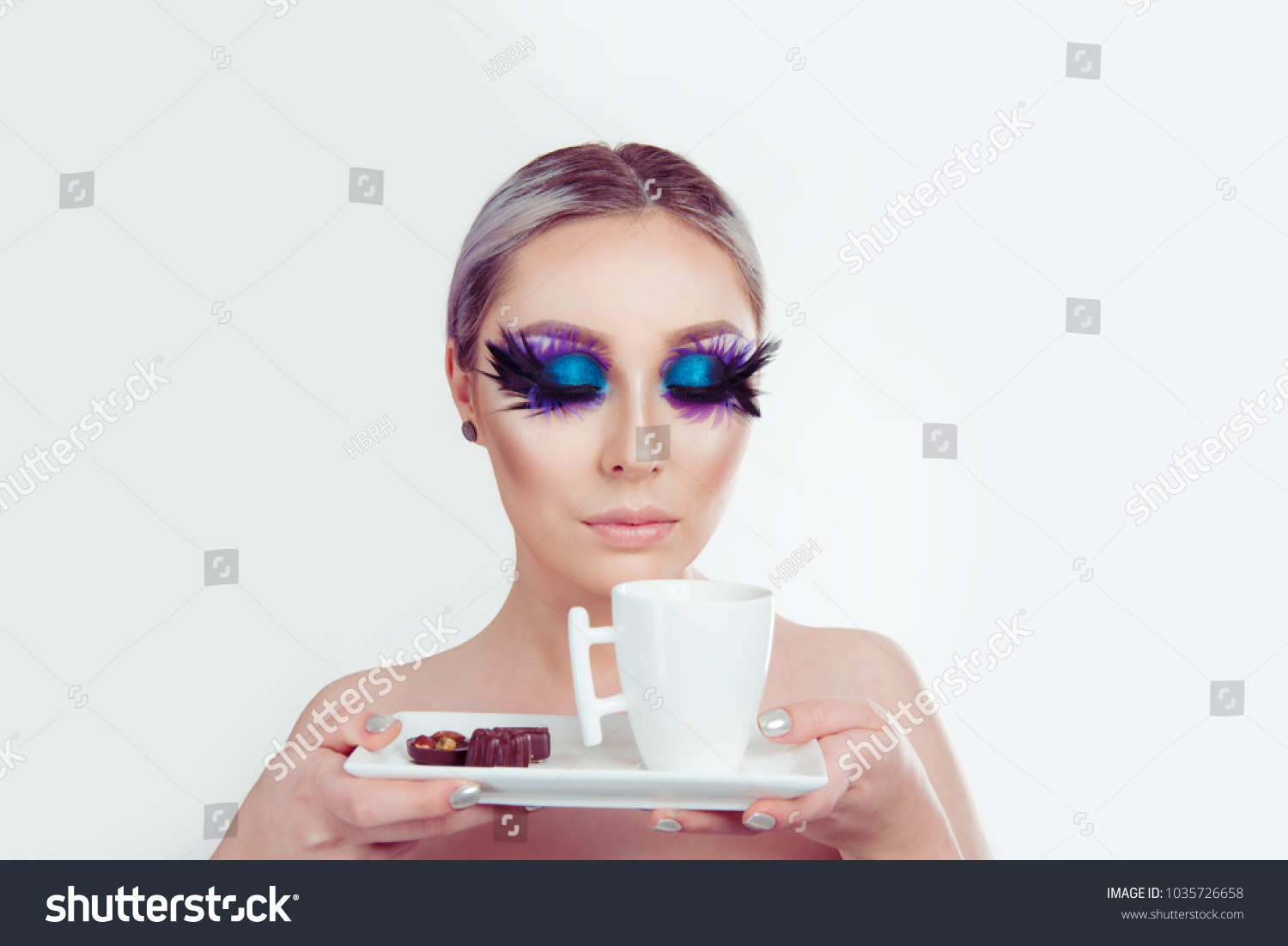 Enjoying aroma of a coffee cup. Beautiful woman with artistic purple blue eyes makeup feather on eyelashes eyes closed holding cup of tea and chocolates on white. Blue black swan and coffee concept #1035726658