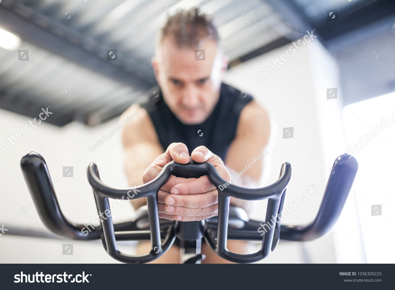 white-haired man in black training indoor cycling in front of window with light holding the handlebar in the middle #1036309225