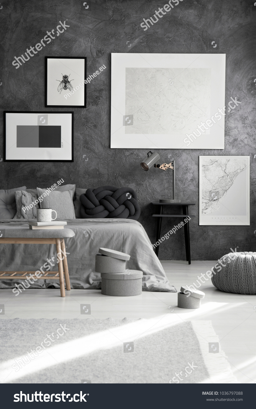 Silver painting and map on a gray wall above bed with dark knot cushion in monochromatic bedroom interior #1036797088