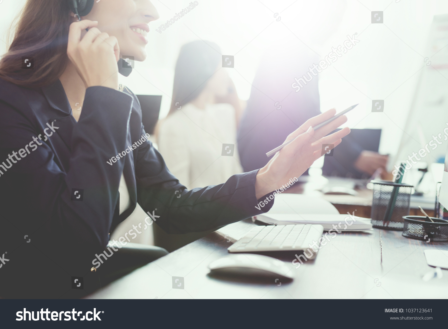 A young girl is looking at a computer screen in the office and is talking on the phone. She works in the call center. She's in a good mood. Call center concept. #1037123641