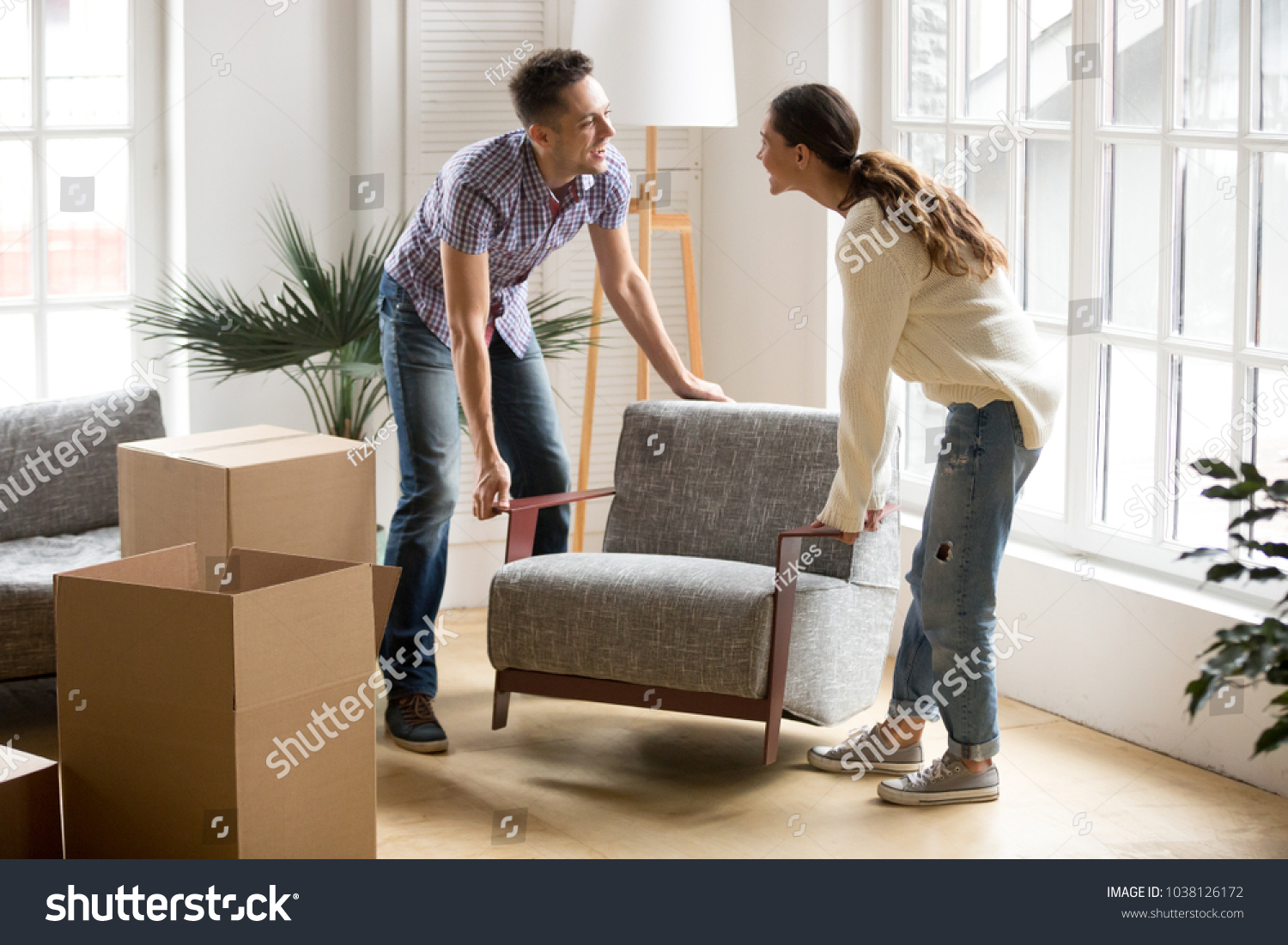 Smiling couple carrying modern chair together placing furniture moving into new home, young family discussing house improvement interior design while furnishing living room, remodeling and renovation #1038126172