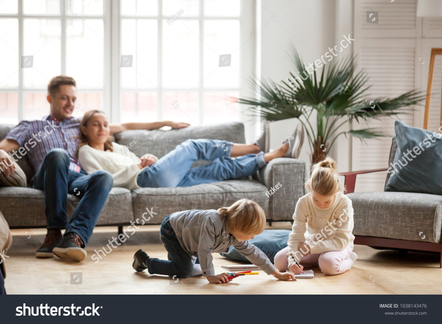 Children sister and brother playing drawing together on floor while young parents relaxing at home on sofa, little boy girl having fun, friendship between siblings, family leisure time in living room #1038143476