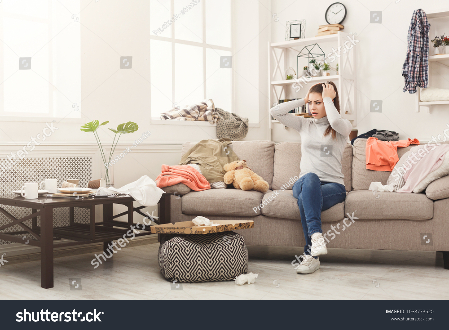 Desperate helpless woman sitting on sofa in messy living room. Young girl surrounded by many stack of clothes. Disorder and mess at home, copy space #1038773620