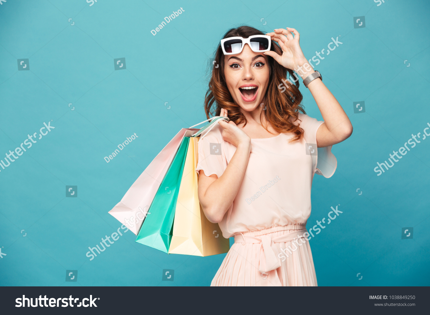 Portrait of an excited beautiful girl wearing dress and sunglasses holding shopping bags isolated over blue background #1038849250