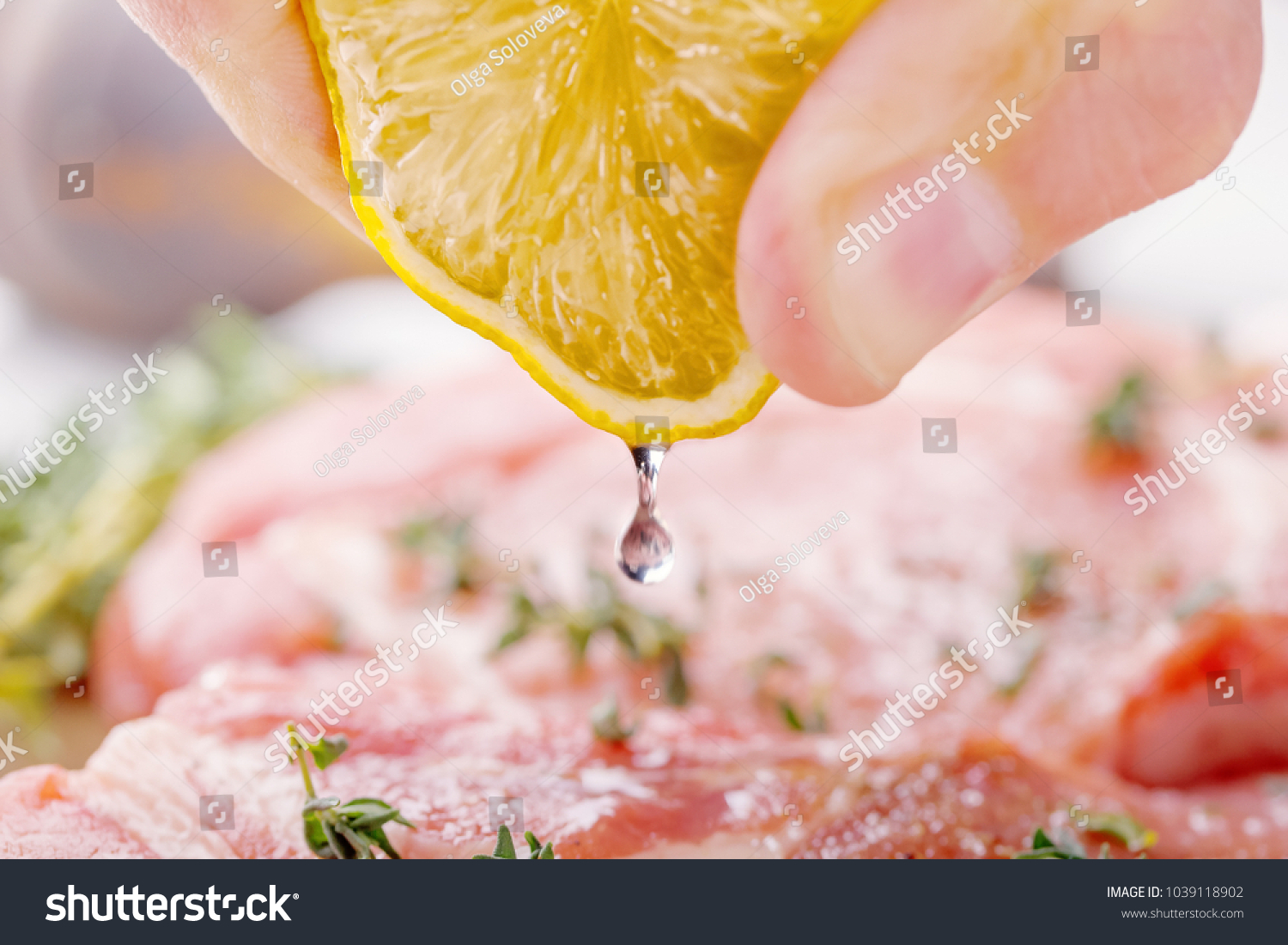 The man's hand squeezes the lemon juice with a raw pork steak. Also on the table, onion, thyme, garlic, salt, pepper, salt and pepper shaker on a wooden cutting board close-up #1039118902