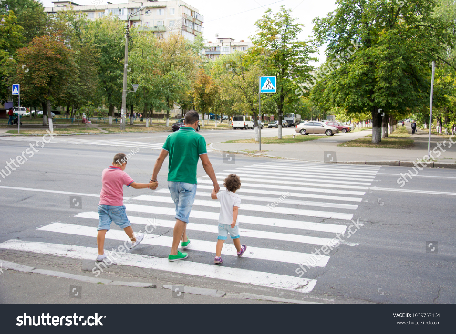 In the summer on the street at the pedestrian crossing father and son and daughter cross the road. Dad holds little hands with his hands. #1039757164