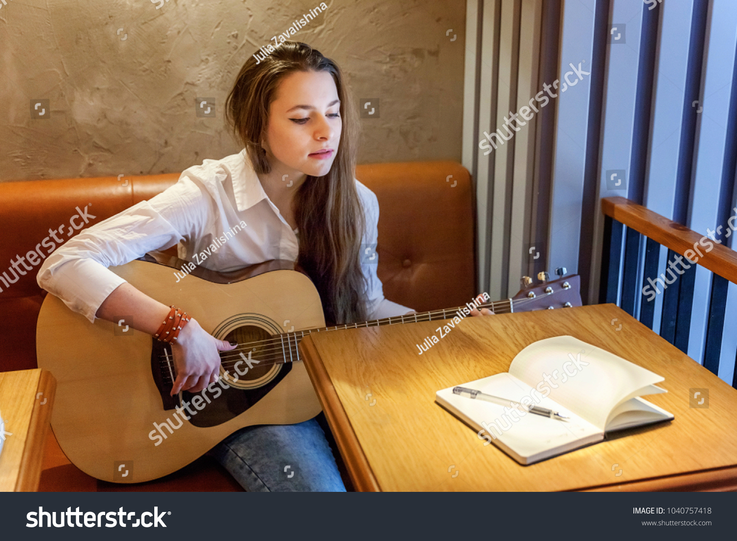 Young smiling hipster woman sitting playing guitar and write a song at cafe. Girl learning to play music #1040757418