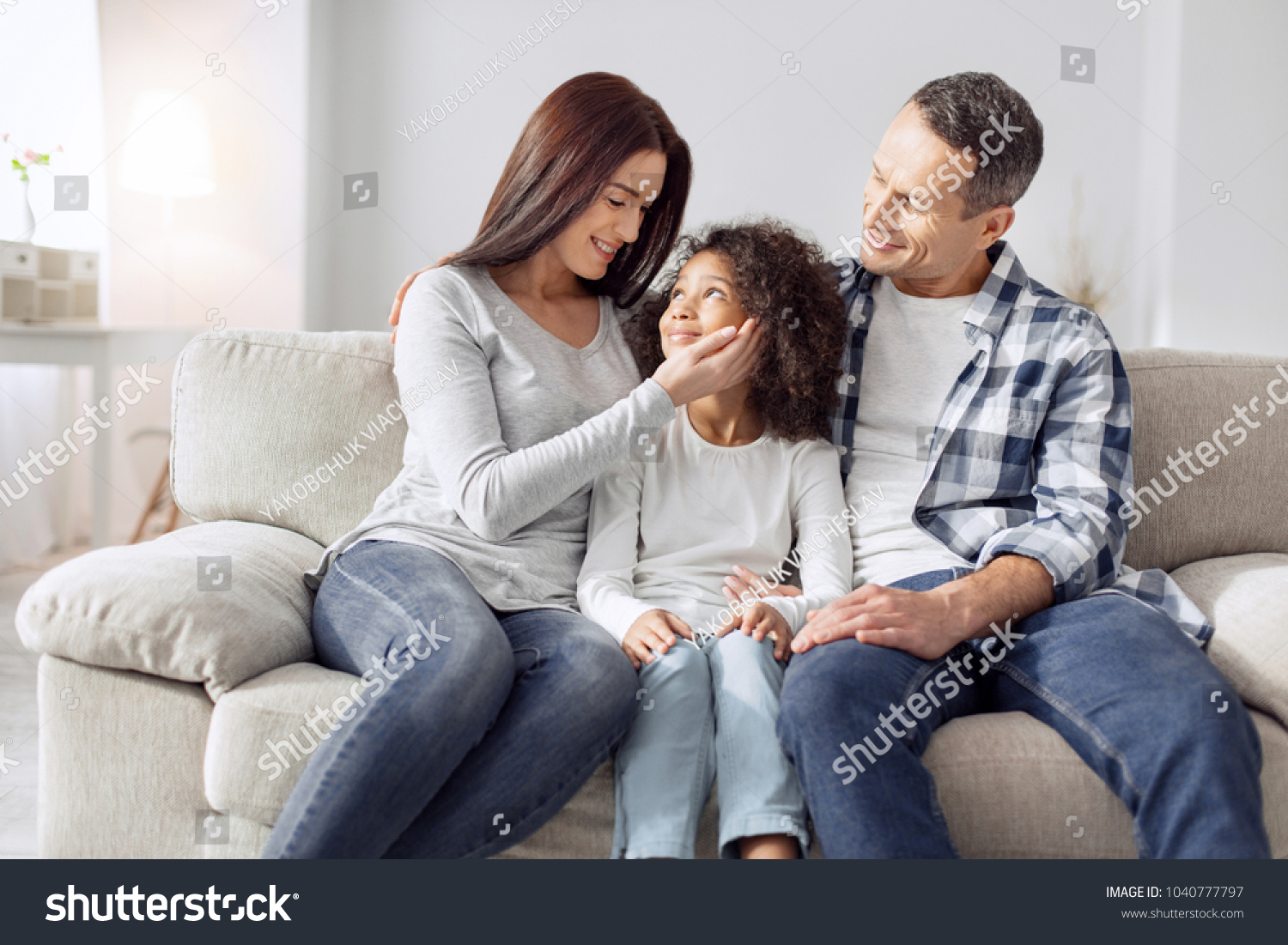 Our treasure. Attractive alert curly-haired girl smiling and sitting on the couch with her parents and they looking at her #1040777797