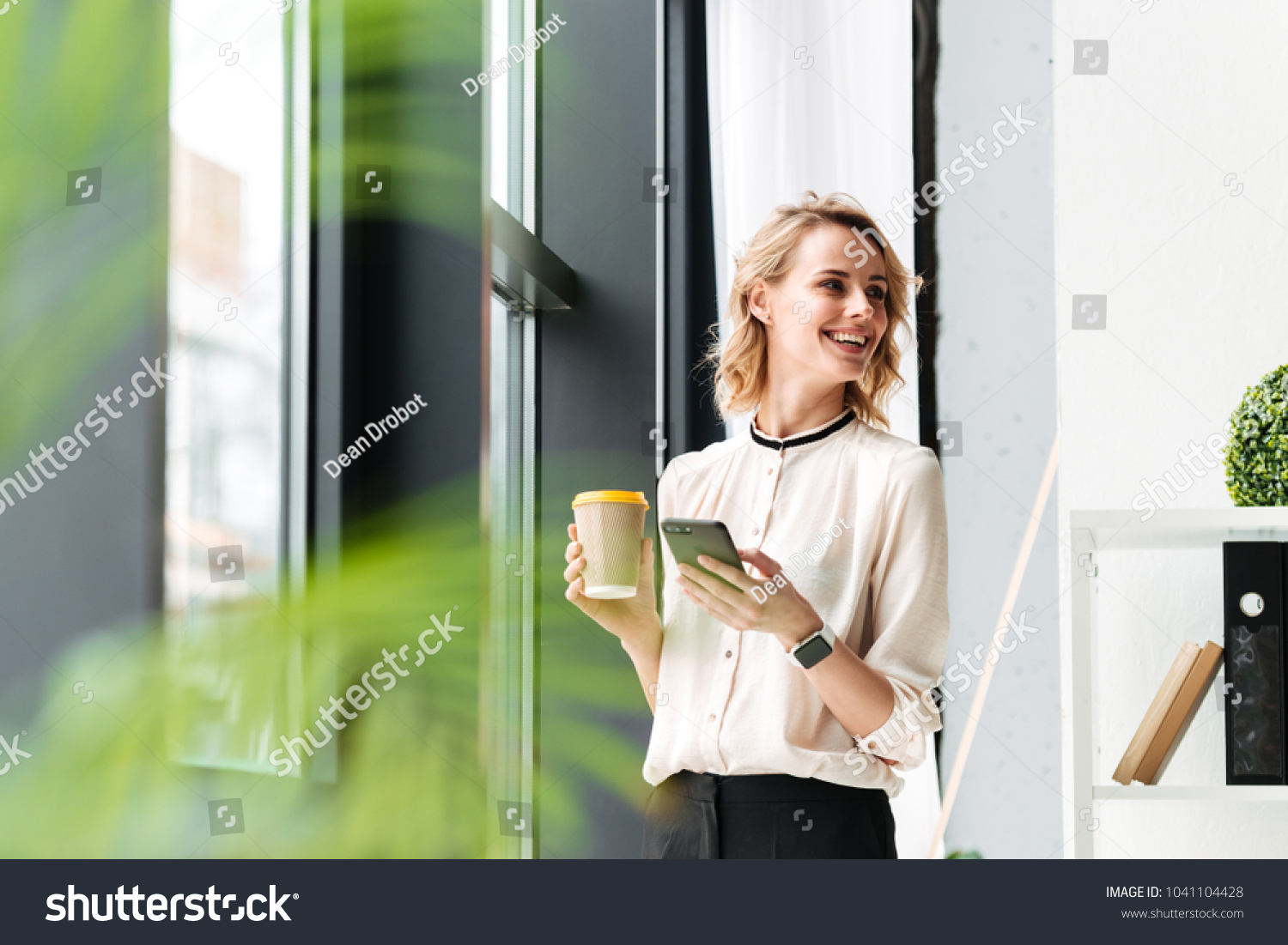 Image of cheerful young business woman in office looking aside chatting by mobile phone drinking coffee. #1041104428