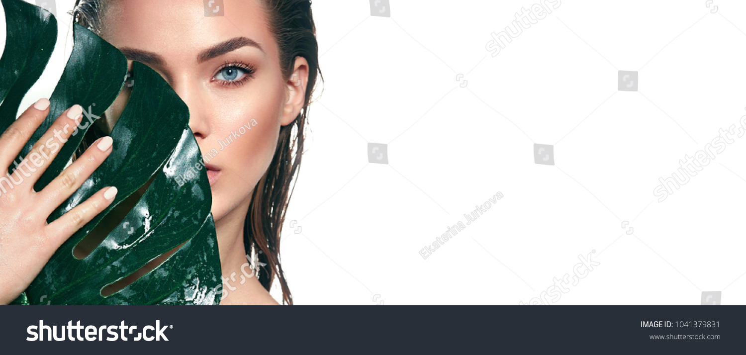 A beautiful young girl in the studio on a white background with wet skin and wet hair holds a large green tropical leaf in hands and covers a part of her face.Fashion, beauty, make-up, cosmetics. #1041379831
