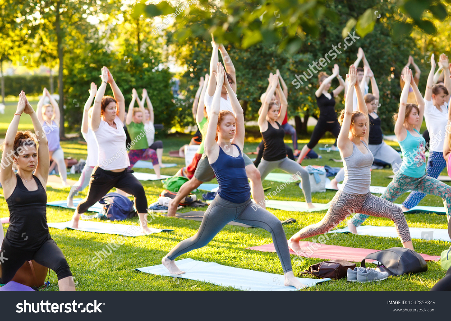 big group of adults attending a yoga class outside in park #1042858849